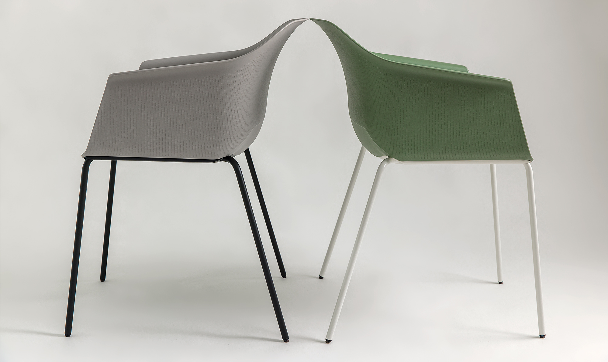 Side view of two Noom 60 chairs designed by Alegre Design for Actiu, positioned back-to-back. One chair is light gray with thin, black metal legs, while the other is olive green with thin, white metal legs. Both chairs feature a curved, bucket-style seat with integrated armrests, emphasizing their sleek and minimalist design.