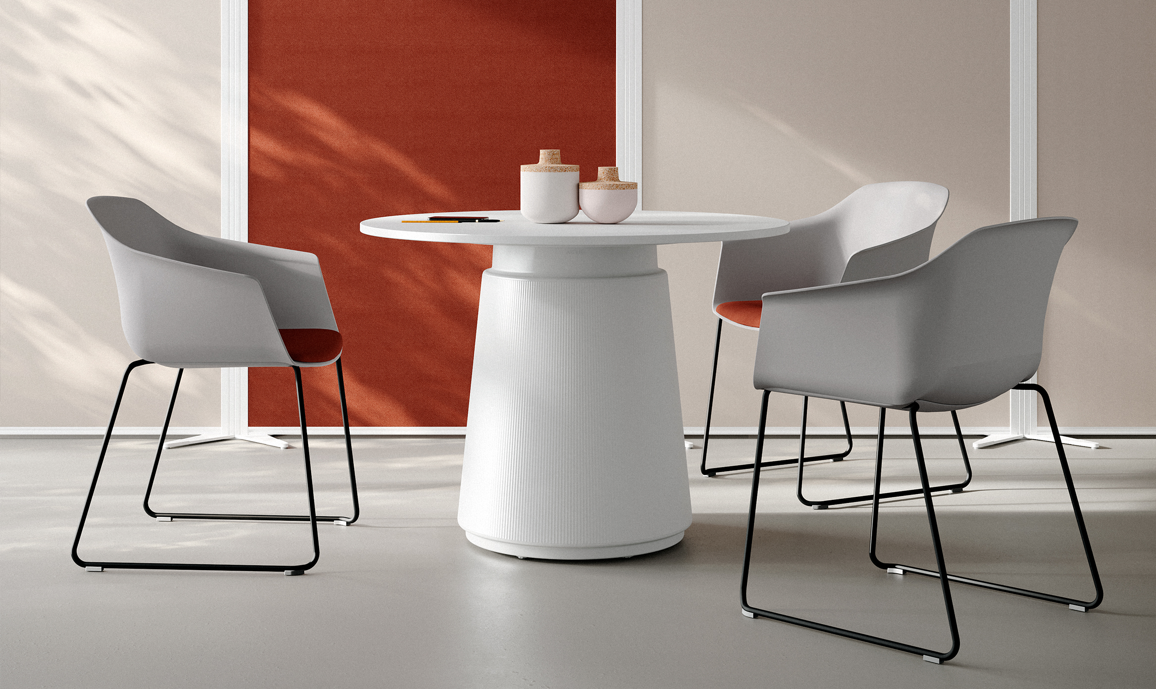 A contemporary meeting space featuring Noom 60 chairs designed by Alegre Design for Actiu. The room includes a round, white table with a cylindrical base, surrounded by four Noom 60 chairs with light gray seats and black metal legs. The chairs have integrated armrests and are accented with orange cushions. The setting is modern and minimalist, with a red and beige wall in the background and soft, natural lighting.