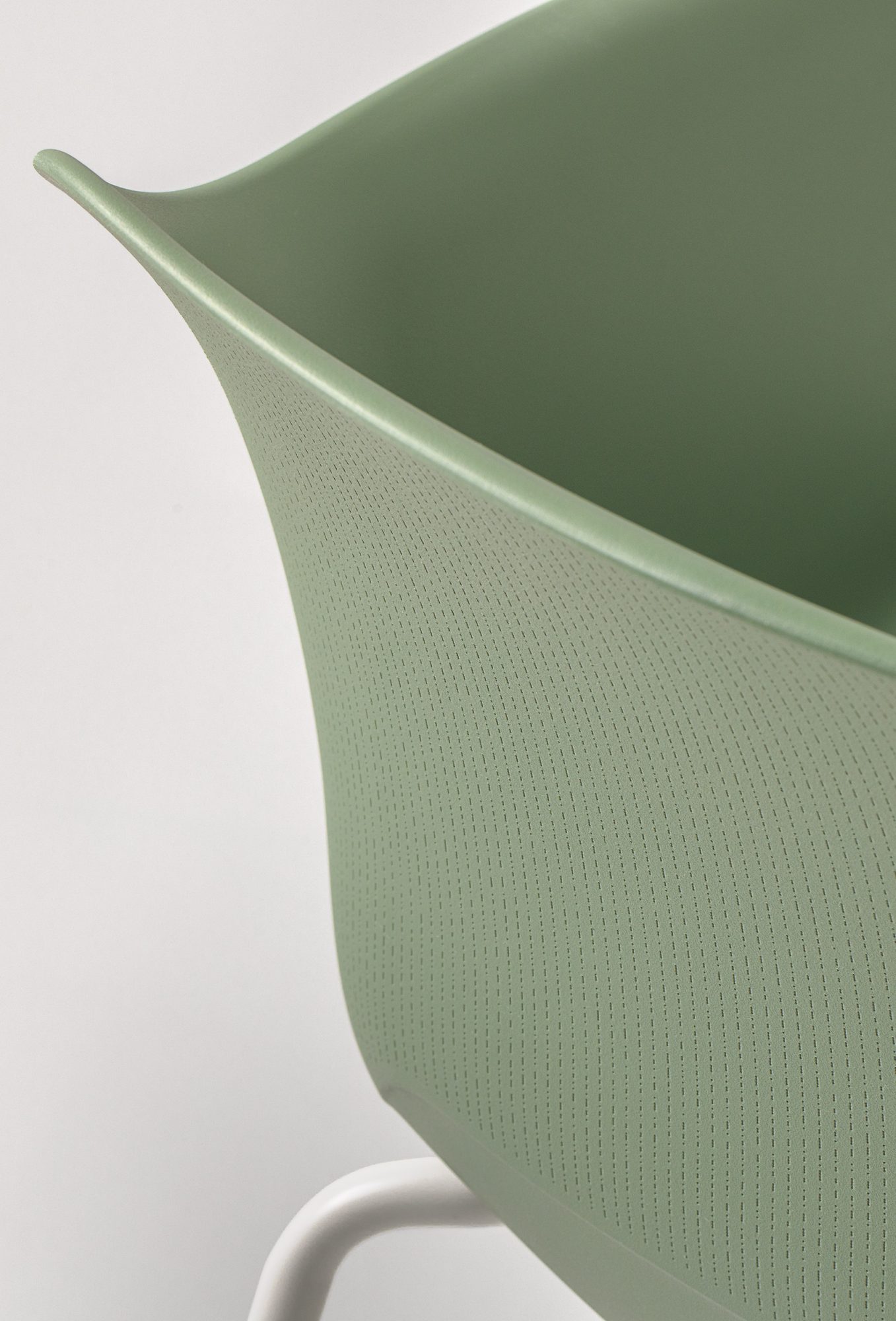 The image shows a detailed close-up of the back texture of the Noom 60 chair designed by Alegre Design for Actiu. The chair features a smooth, green surface with a subtle, fine-textured pattern. The white metal legs are partially visible, emphasizing the chair's minimalist and modern design.