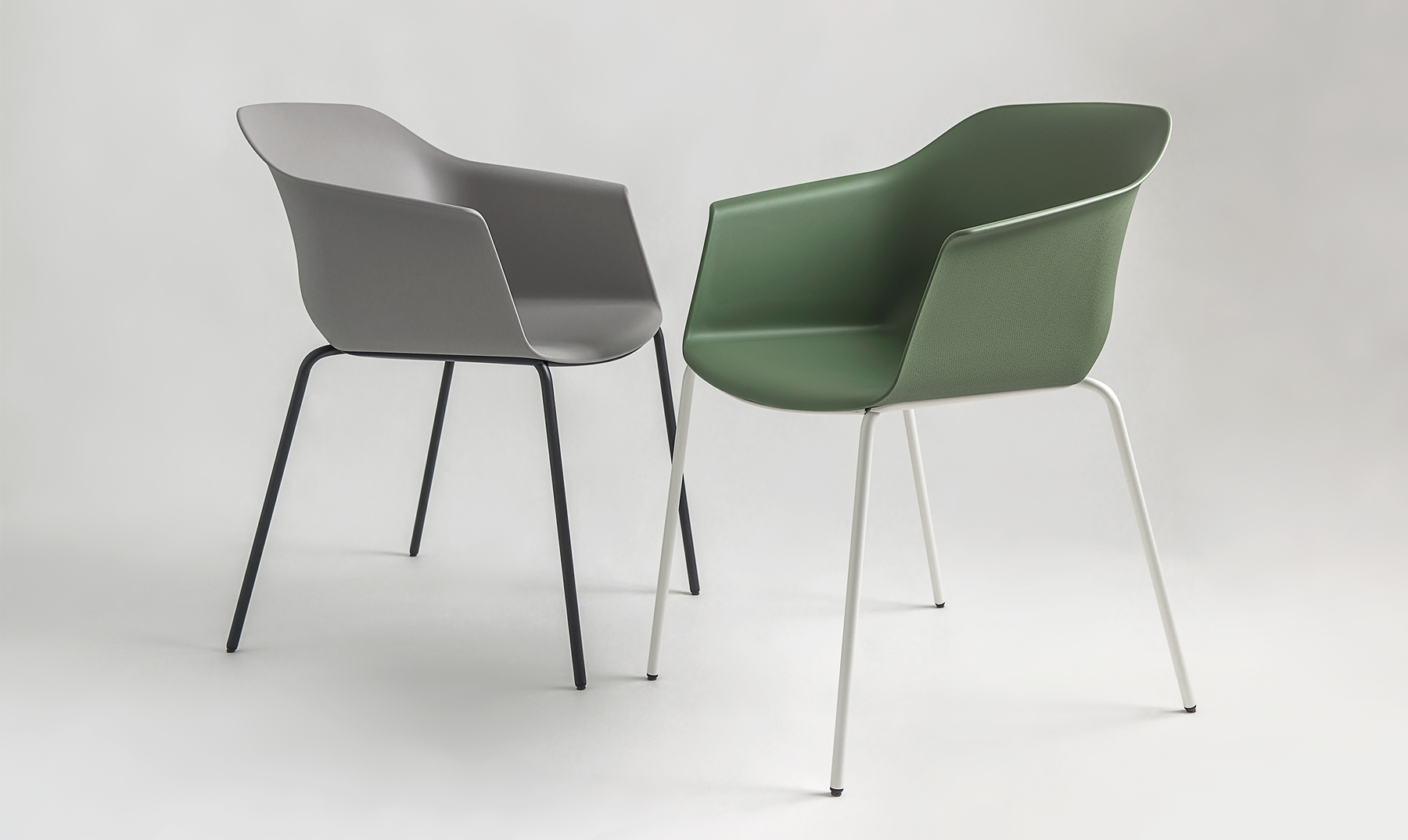 The image shows two modern, minimalist chairs named Noom 60 designed by Alegre Design for Actiu. One chair is light gray with thin, black metal legs, while the other is olive green with thin, white metal legs. Both chairs feature a curved, bucket-style seat with integrated armrests. These sleek and contemporary chairs are suitable for various settings such as offices or homes. Keywords: Noom 60, Actiu, Alegre Design, product designer, modern chairs, minimalist chairs, office furniture, contemporary design.