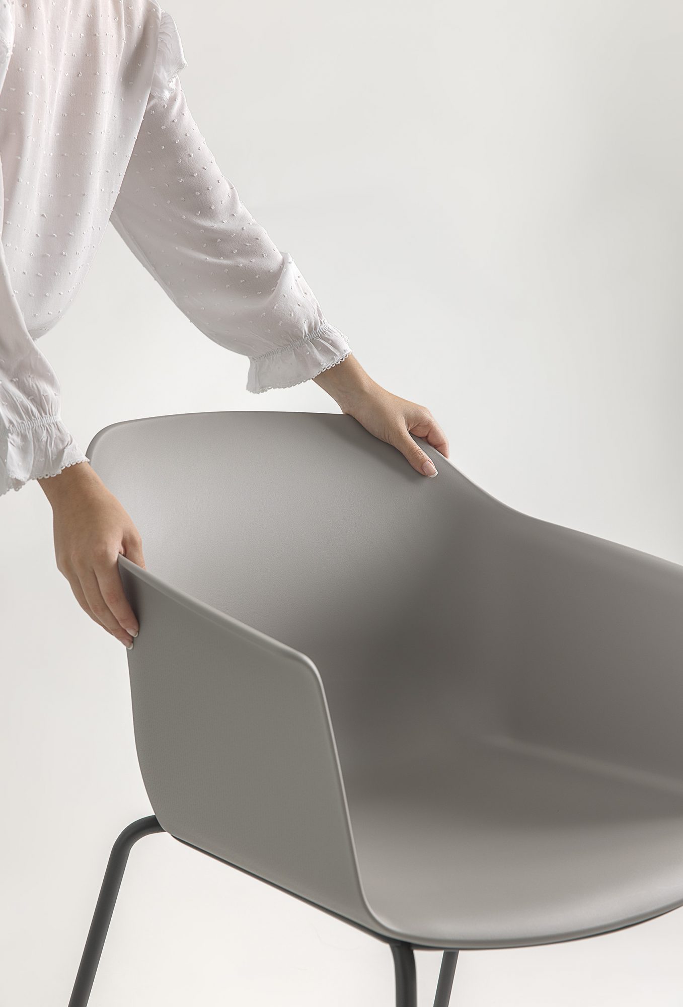 A person adjusting the Noom 60 chair designed by Alegre Design for Actiu. The chair has a smooth, light gray, curved bucket-style seat with integrated armrests and black metal legs. The person is holding the chair from the back and side, highlighting its ergonomic design and ease of handling.